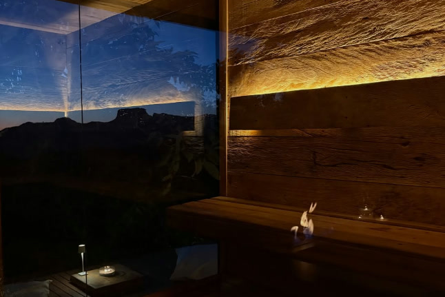 Interior da sauna em madeira, com luz quente embutida e vista da serra ao crepúsculo através do vidro