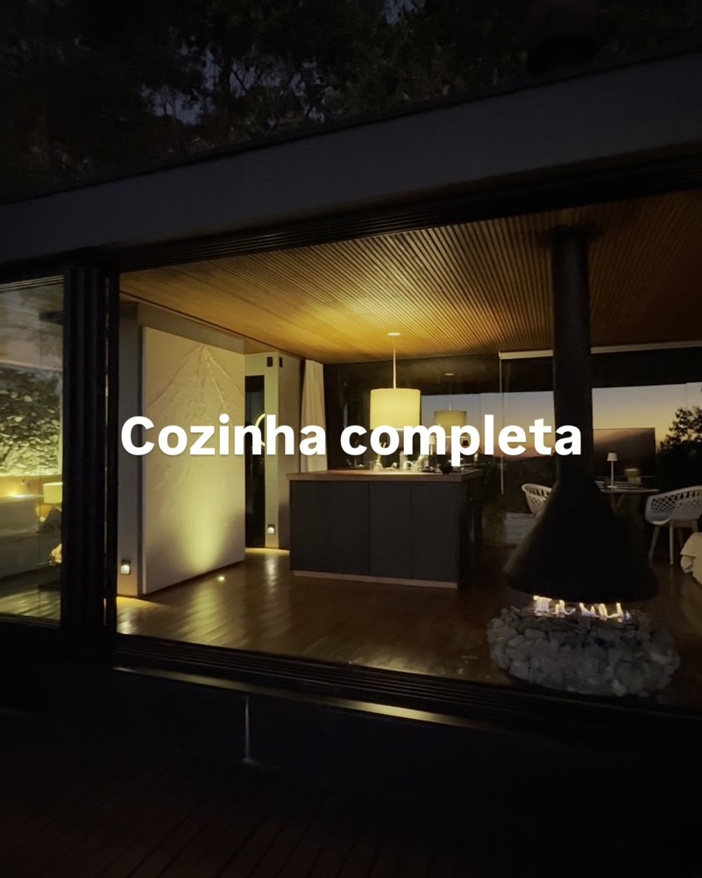 Interior da casa iluminado à noite, com paredes de vidro para a mata