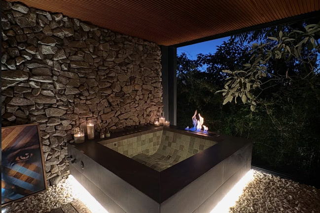 Jacuzzi quadrada à noite, com velas acesas, lareira e parede de pedra