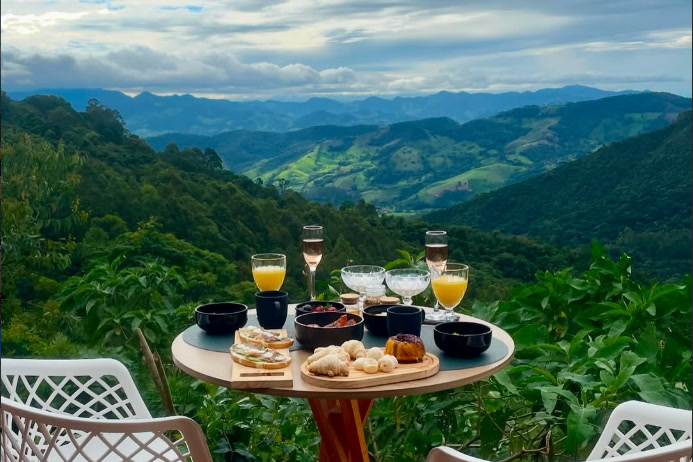 Mesa de café da manhã no deck, duas cadeiras brancas, vale e serra ao fundo em dia nublado